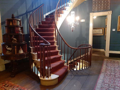 The interior of Houmas House is stately and beautiful with a wide center hall with large portraits on the wall. The second photo shows a fireplace and artwork; the third photo shows a desk in a stately room with a portrait in an ornate frame. The last photos are of bedrooms and a curving staircase.