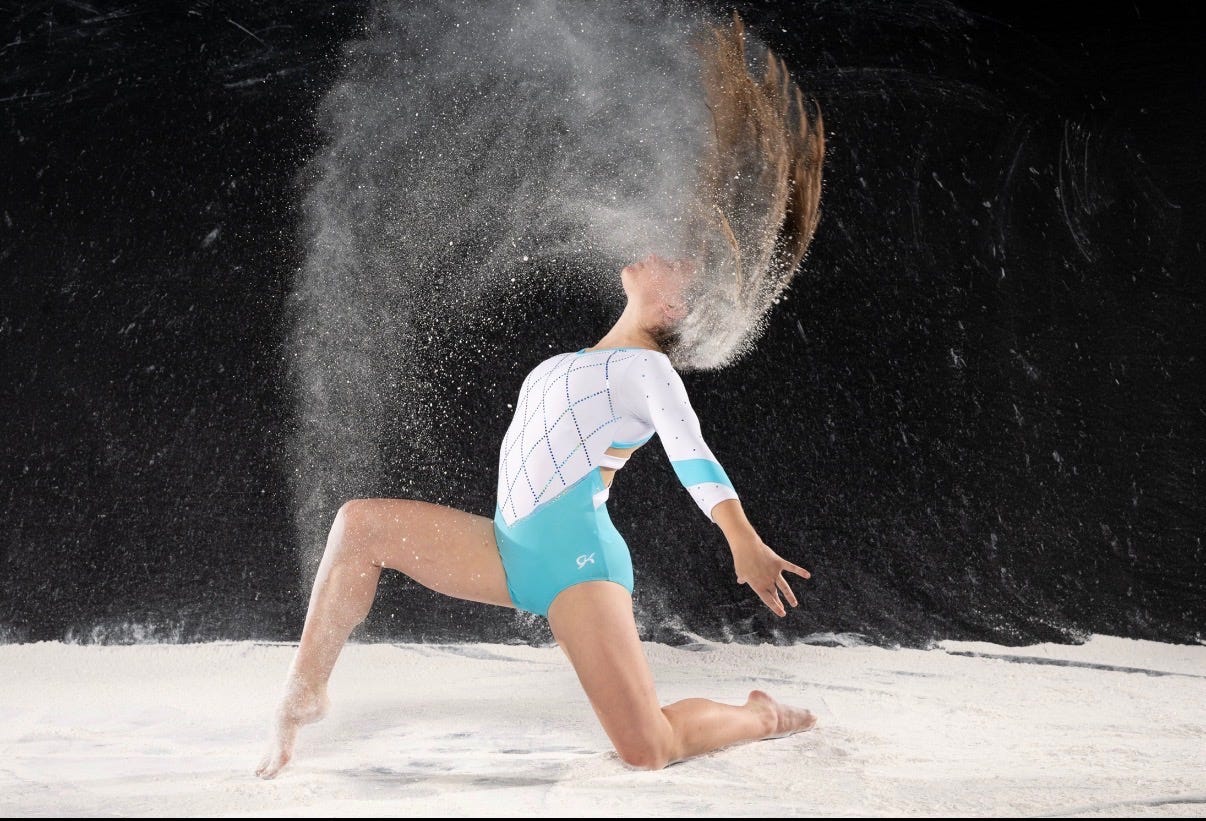 A cinematic beauty shot of The Beauty Ed® THE LAB mentee Lexie Stone in a teal gymnastics leotard, kneeling on a dark floor surrounded by a cloud of white gymnastics chalk (magnesium carbonate).