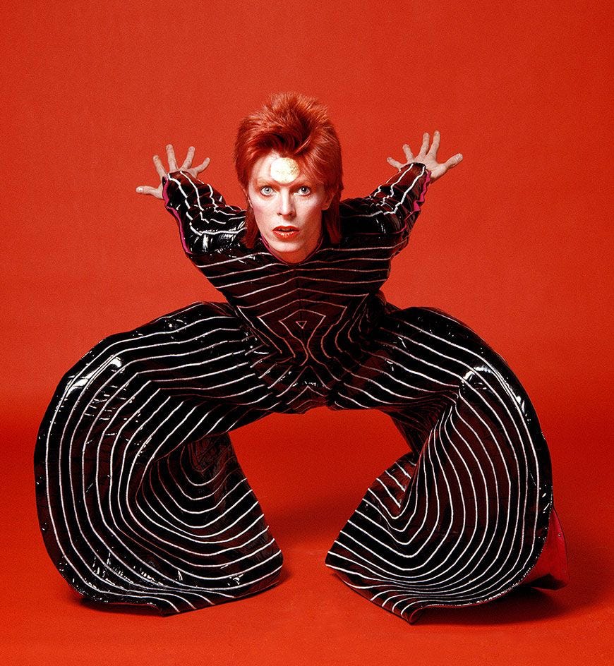 David Bowie as Ziggy Stardust in black striped jumpsuit with red hair and lightning makeup, iconic glam rock studio portrait against red background.