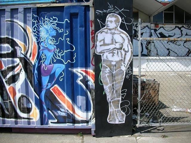 Street art on a metal panel: a blue-toned mythical woman with swirling hair and a shirtless male figure on a black strip, set beside a chain-link fence. Jeremy's artwork is of the shirtless man, wearing black pants and boots. He is muscular, and he has his head turned to the side. His right arm is crossed over his body, and his left hand is in his pocket. This artwork resembles a Tom of Finland artwork. Street art on a metal panel: a blue-toned mythical woman with swirling hair and a shirtless male figure on a black strip, set beside a chain-link fence. Jeremy's artwork is of the shirtless man, wearing black pants and boots. He is muscular, and he has his head turned to the side. His right arm is crossed over his body, and his left hand is in his pocket. This artwork resembles a Tom of Finland artwork.
