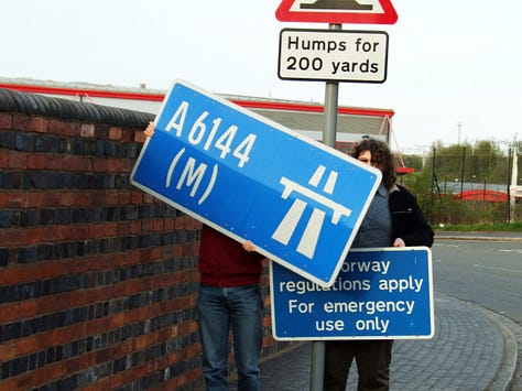 Three photographs showing people posing with the A6144(M) sign in various settings - on the left in front of a building; in the middle, two people obscured by the signs and clearly having trouble holding them; on the right a whole group posing together with the sign in a lay-by