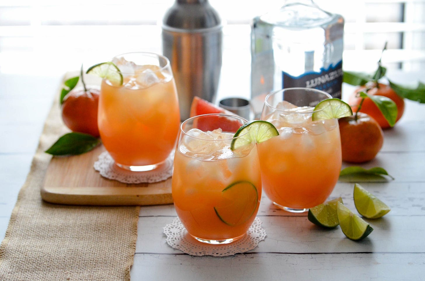 three glasses of a citrusy cocktail called a calamity jane on a wood background with lime wedges and oranges. 
