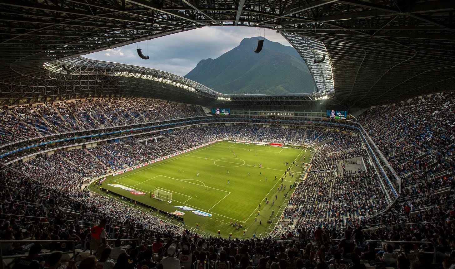 High-angle view inside a packed modern soccer stadium during a match, featuring an open roof structure framing a scenic mountain in the background.