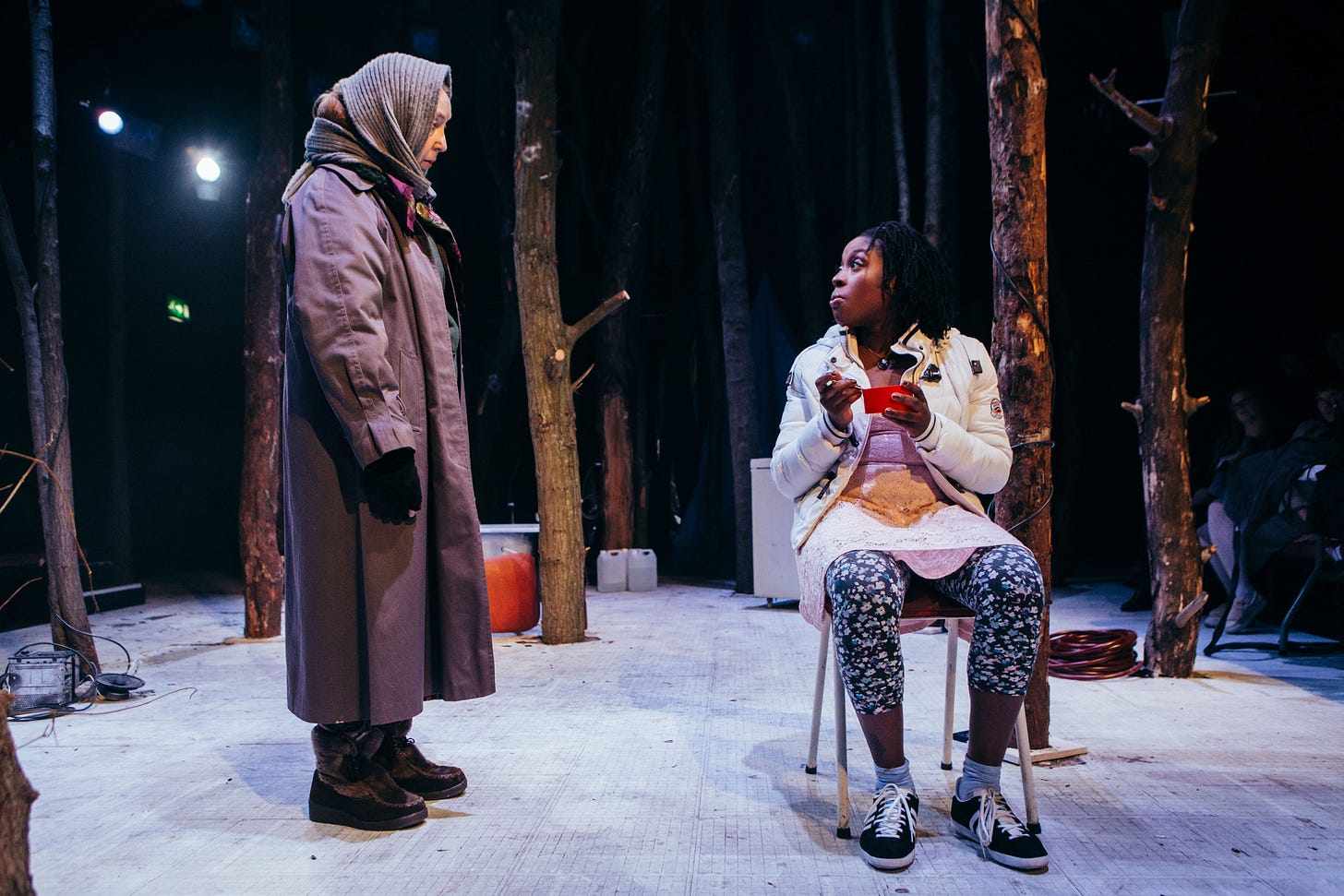 A woman in a winter coat and headscarf talks to a younger woman seated on a chair in the snow.