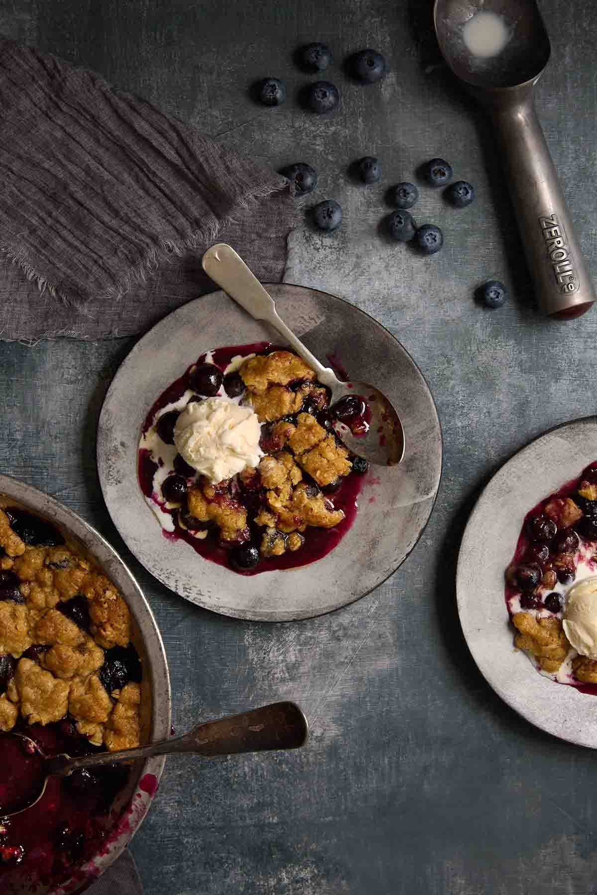 A freshly baked blueberry crumble in a baking dish with a golden, oat-based topping and a bubbling purple berry filling.