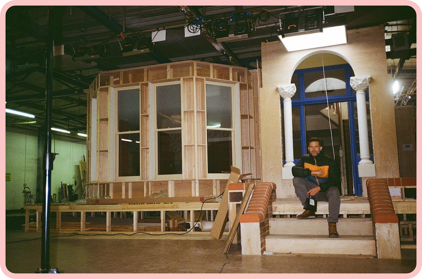 A photo of the wooden exterior of a Victorian-style house which is still in the process of being constructed. Three large steps lead to the front door, and a white man in a blue and orange jacket is sitting on them and staring directly at the camera. The wooden flooring, industrial lighting, and warehouse-style ceiling, suggest that the house is not in fact outside, but indoors, in a film studio. 