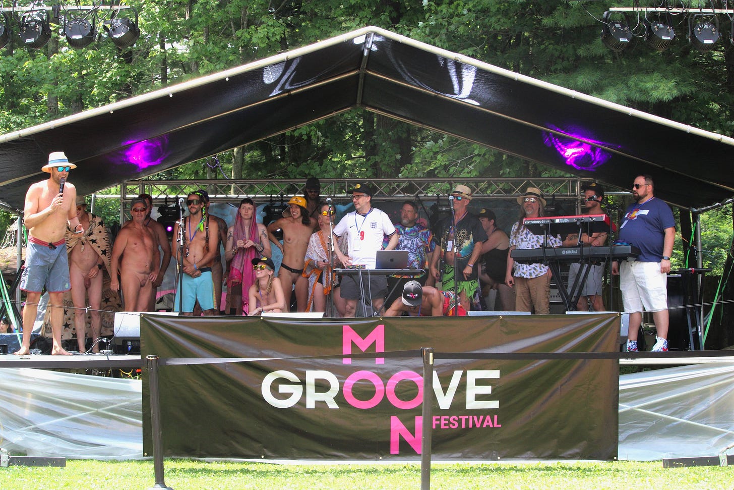 Eli Porr speaks into a microphone from the edge of the main stage while a large group of MoonGroove staff and volunteers stand together behind him for a recognition moment, with the MoonGroove banner visible below the stage. Eli Porr speaks into a microphone from the edge of the main stage while a large group of MoonGroove staff and volunteers stand together behind him for a recognition moment, with the MoonGroove banner visible below the stage.
