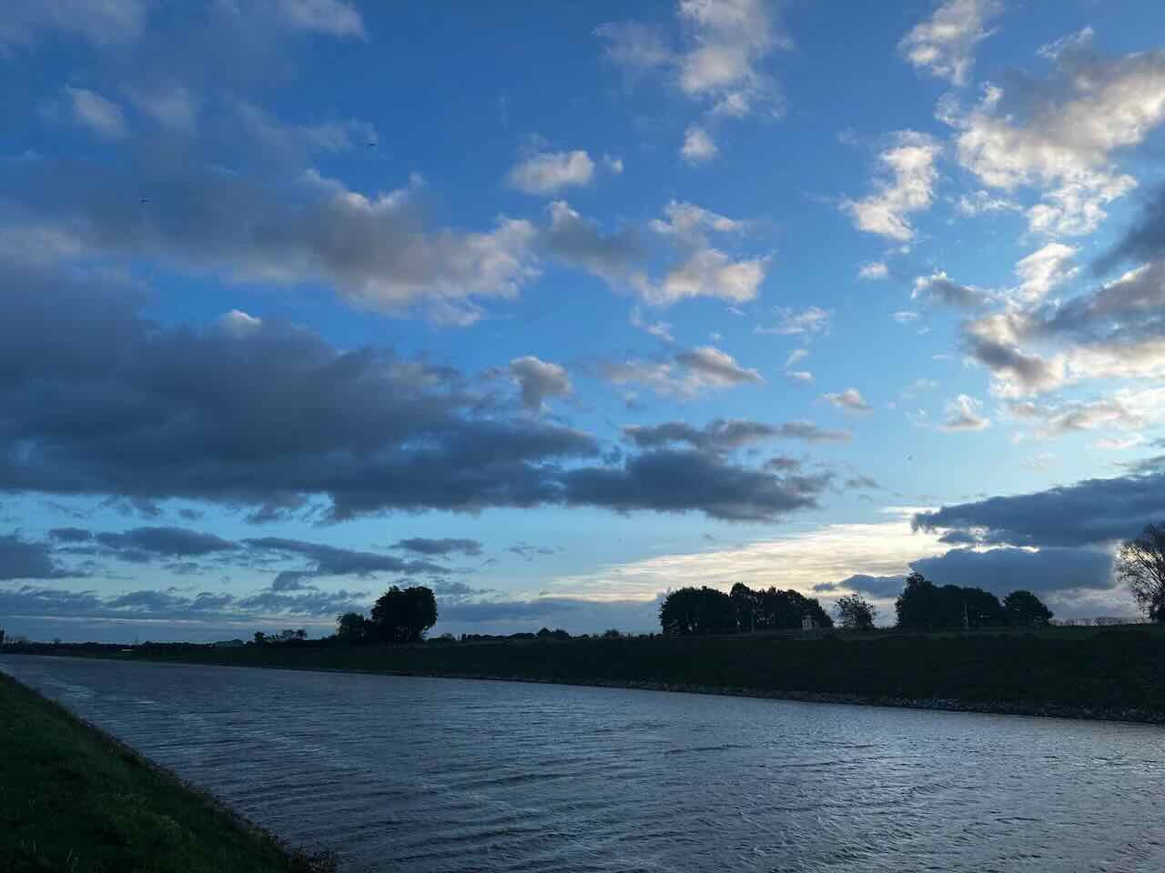 A calm early morning scene showing the Julianakanaal in the south of the Netherlands, with soft blue skies, scattered clouds, and gentle light reflecting on the rippling water.