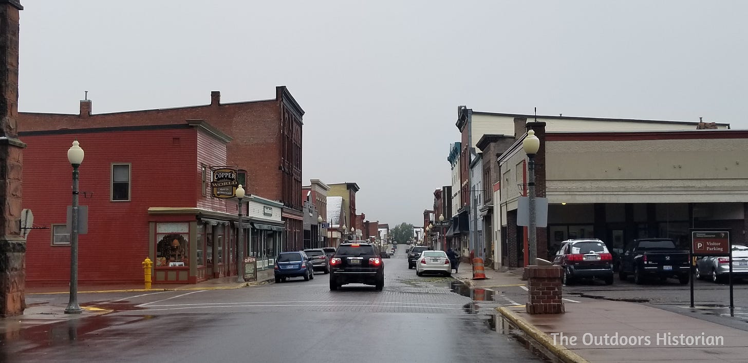 A daytime view looking north up 5th Street in downtown Calumet during the fall. The historic brick buildings line the street, showing the town's resilient architecture and quiet commercial life.