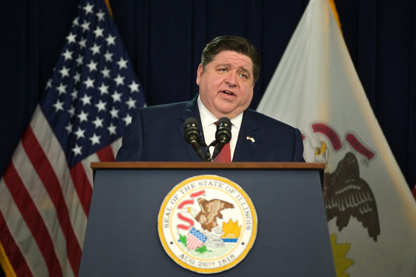 Pritzker speaking at a podium with the IL seal on it; us and IL flags behind him