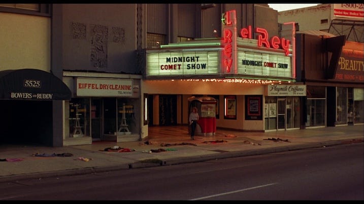 Night of the Comet (1984) promotional still of a woman approaching the El Rey Theater under a marquee that reads “Midnight Comet Show,” surrounded by piles of clothing.
