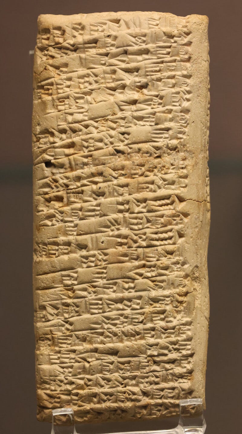 A tall clay tablet standing on small clear mounts against a plain brown museum wall, its surface packed edge to edge with rows of cuneiform pressed into the wet clay. A tall clay tablet standing on small clear mounts against a plain brown museum wall, its surface packed edge to edge with rows of cuneiform pressed into the wet clay.