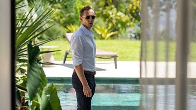 A man stands by a swimming pool, wearing a shirt and trousers and a pair of dark sunglasses