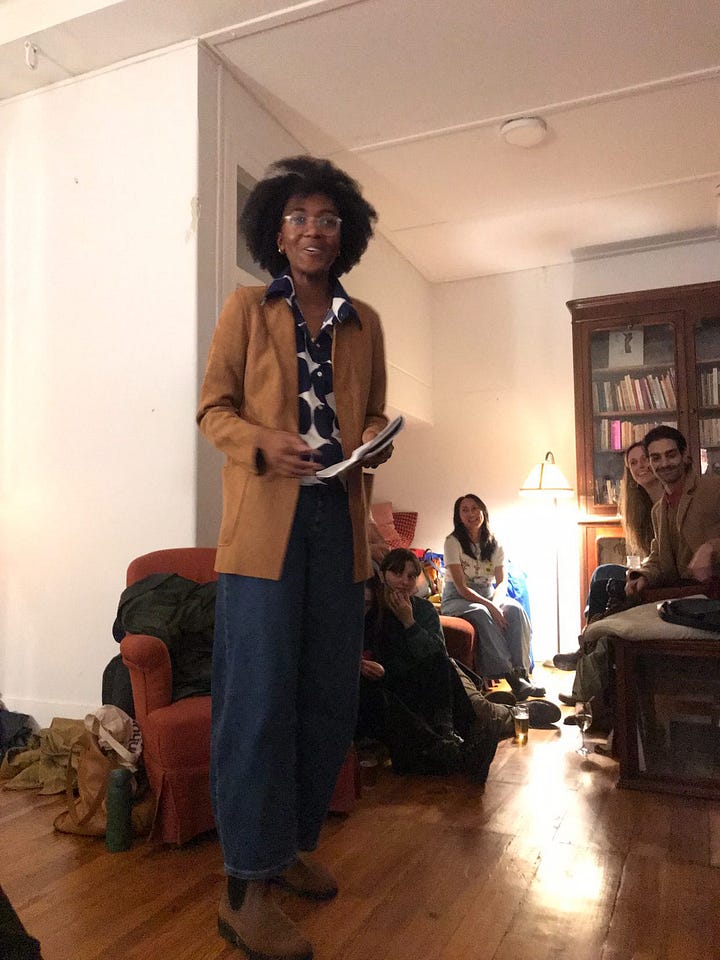 photos of monét standing while talking to the crowd and sitting listening to the tidbit of the episode. she's wearing a blue and white polka dot shirt, a camel jacket, and blue jeans with white nike socks and brown boots.