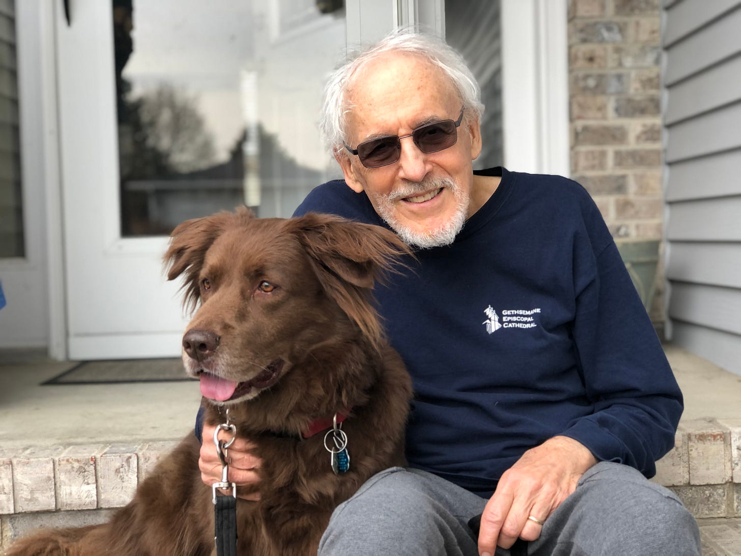 A smiling white-haired man in sweats and sunglasses sits outside on the steps with his arm around a big, brown dog