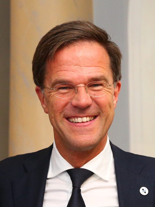 head shot of Mark Rutte smiling. Pale skin, brown short hair, black jacket, white collared shirt and black tie.