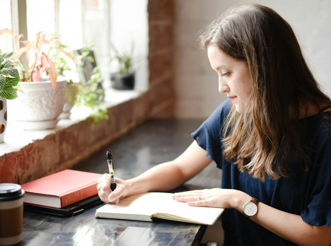 woman sitting in front of black table writing on white book near window