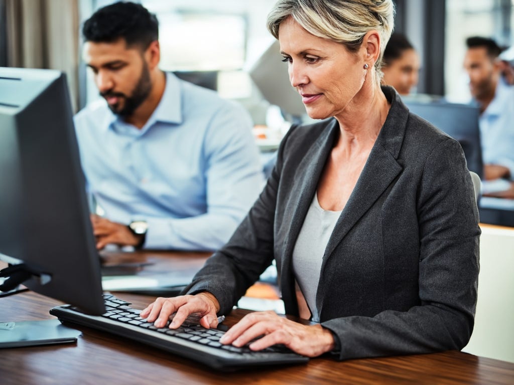 White woman with dishwater blonde hair works intently at a keyboard and monitor. Three brown-skinned employees, as if working in a newsroom, are in the background, slightly blurred.