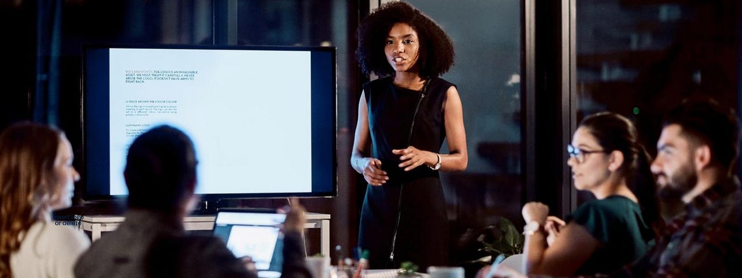A woman standing and giving a presentation to a group of people in an office setting, with a screen displaying text behind her. A woman standing and giving a presentation to a group of people in an office setting, with a screen displaying text behind her.