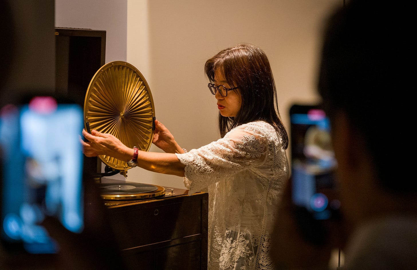 Bystanders record videos as a woman dressed in a flowing white shirt maneuvers a pleated golden disc into position over a shellac record