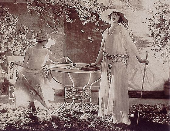 A black and white photograph showing two elegant women, one sitting at a garden table, the other standing next to it. Fashion study two models at a table 1920 by Adolf de Meyer 