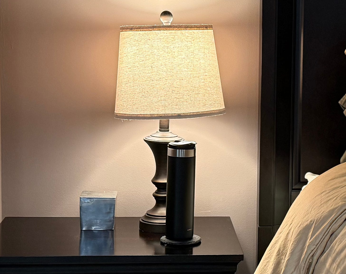 A nightstand in warm evening light — a thermos of hot water beside a lamp, the bed already turned down. The quiet before sleep that most women have to decide to take.
