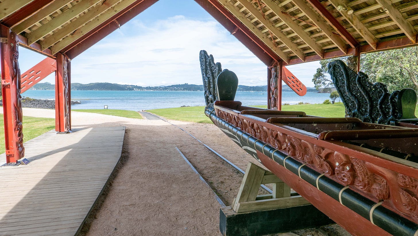 One of the war canoes at the Waitangi Treaty House, under a shelter and with the blue bay in the background.