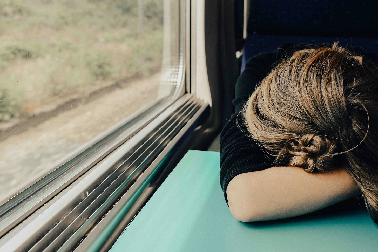A woman with brown hair pulled up in a bun is sleeping on a train with her head face down on her arm.