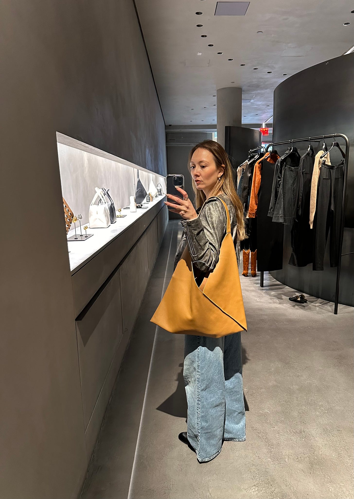 Mirror photo of a woman in denim and a leather jacket holding a large camel tote in Khaite’s SoHo store.
