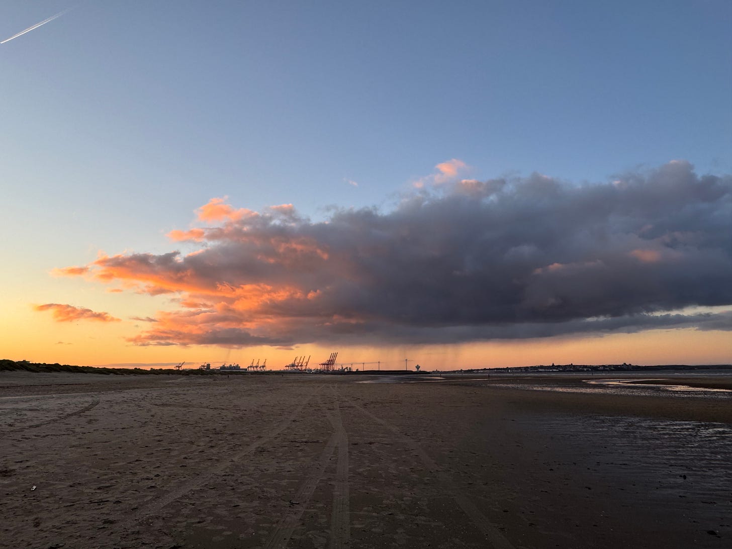 Crosby Beach at sunrise, The cranes at Liverpool Docks can be seen in the distance, silhouetted against the yellow and blue sky, with clouds overhead.