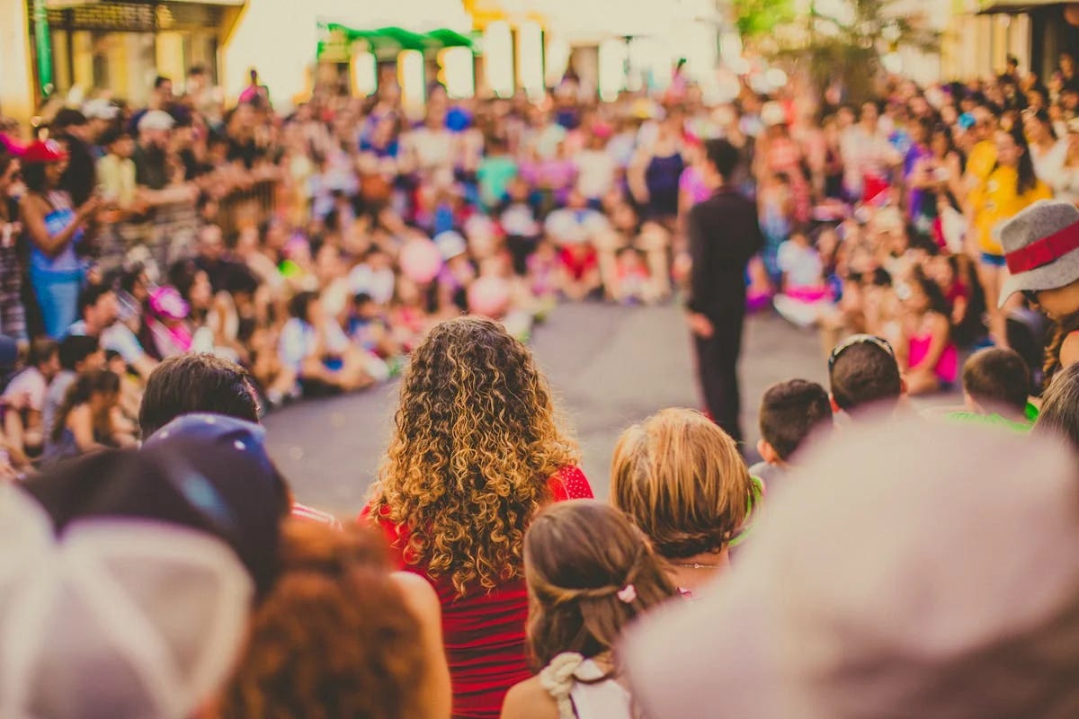 Crowd watching a performance from behind, showing backs of heads with a person in a red striped shirt with curly hair in focus, and a blurred audience in the background with warm lighting and festive atmosphere.
