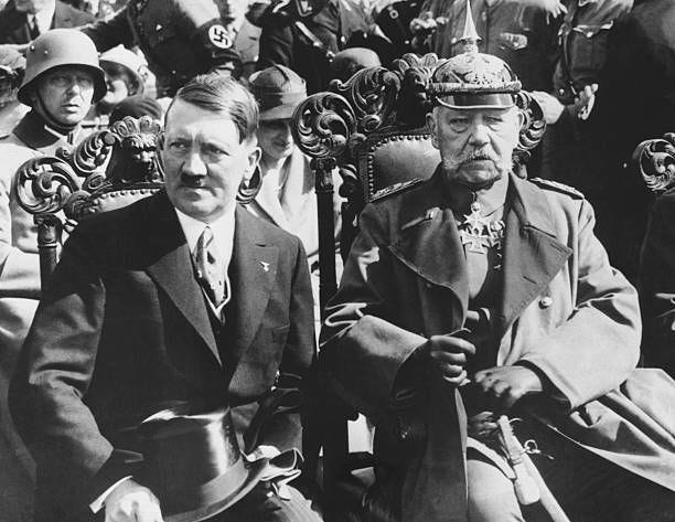 Adolf Hitler and Paul Von Hindenburg sit on ornate chairs surrounded by Nazi soldiers. Von Hindenburg opposed Nazi politics but was forced to appoint...