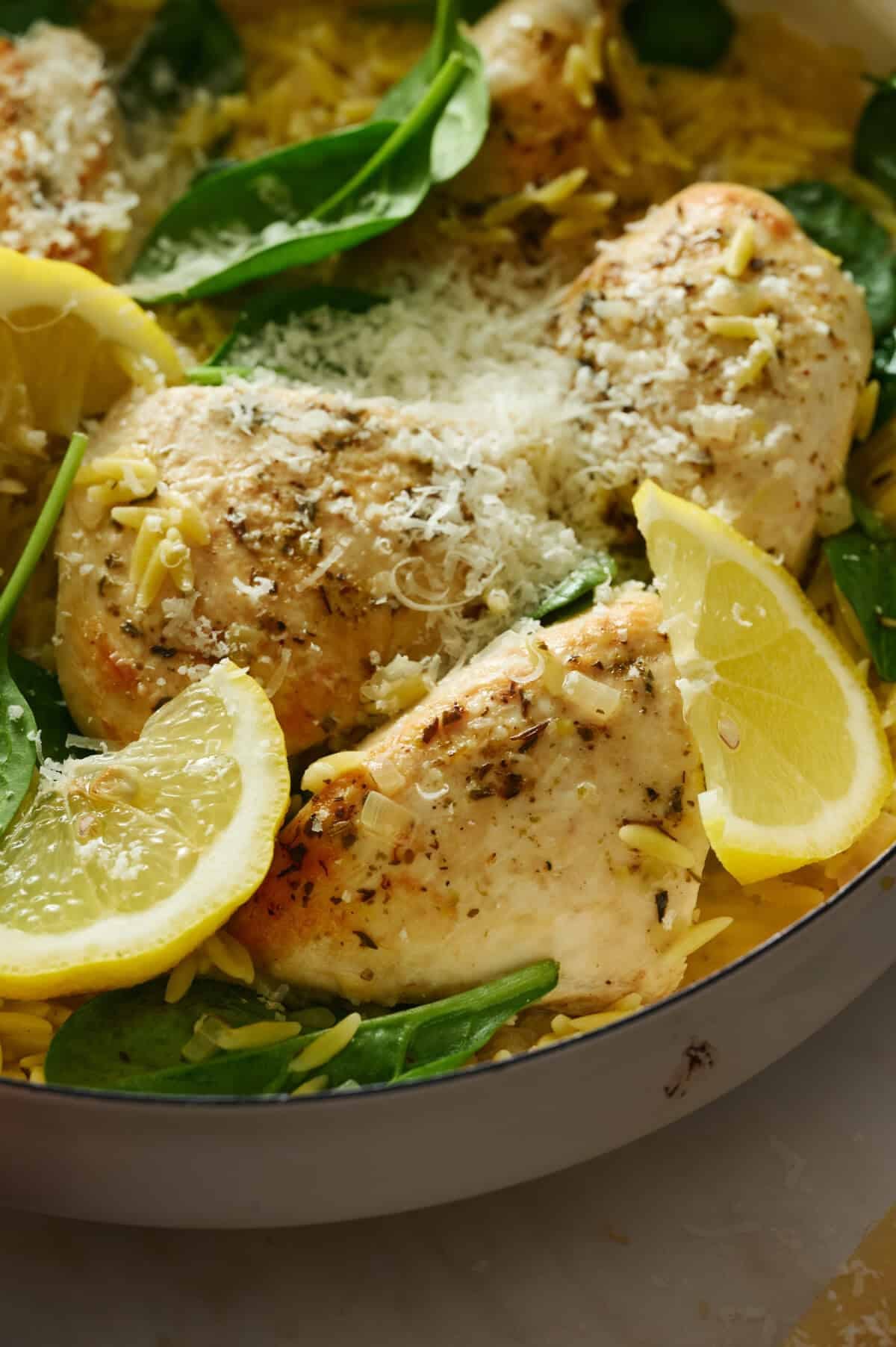 Lemon chicken orzo pasta in a white pot on a marble table with shredded parmesan cheese and bright green spinach leaves.