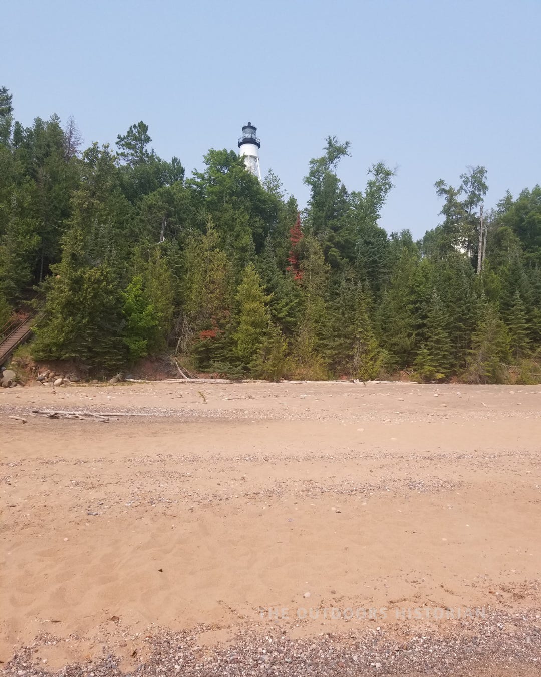 A vertical (4:5) photo of the sleek, white modern Michigan Island Lighthouse in the Apostle Islands. The lighthouse tower rises from a vibrant green tree line behind a strip of sandy beach and blue Lake Superior water.
