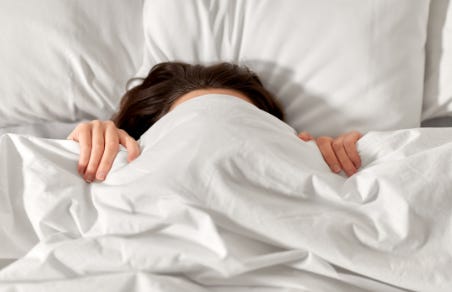 A person in bed with the photo only showing their upper body, hidden underneath a white comforter, with only the top of their head and hands that are holding up the blanket showing.