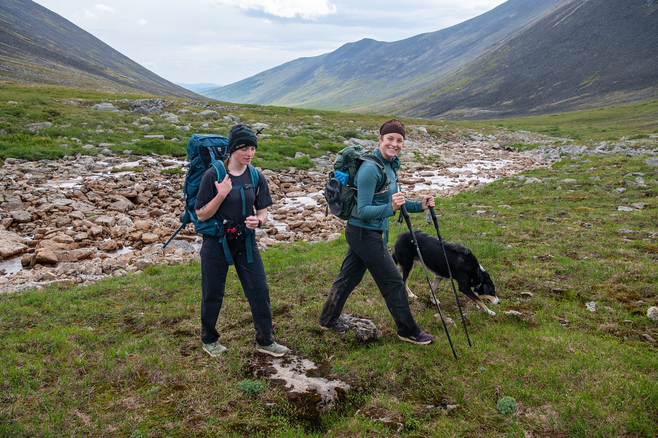 Two hikers with packs and a black-and-white dog walk across a green U-shaped valley floor scattered with pale boulders and threads of shallow water, one hiker smiling toward the camera. Grassy slopes climb to bare, rounded ridges on both sides under a flat grey sky.