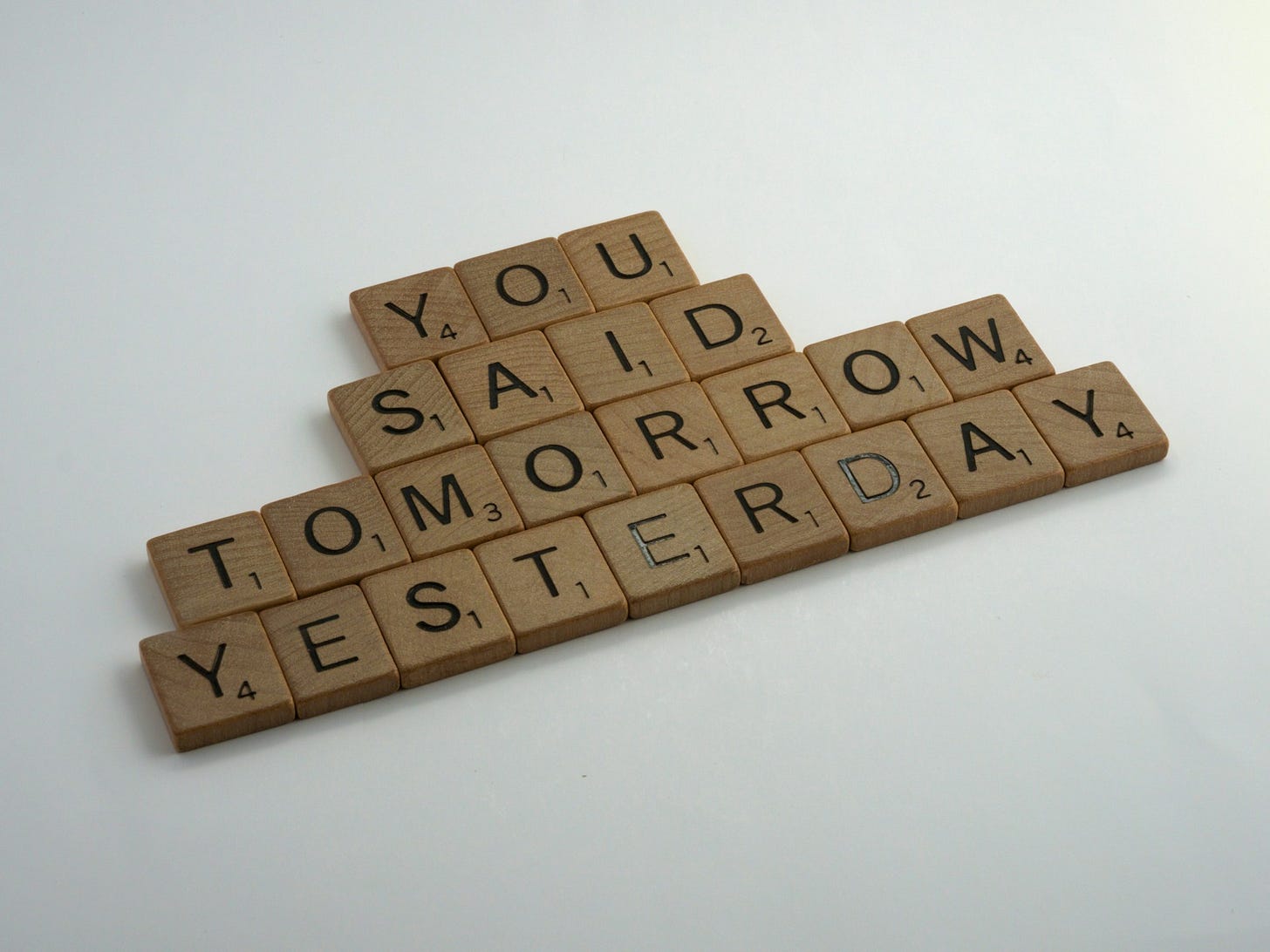 Wooden Scrabble tiles arranged to spell out "YOU SAID TOMORROW YESTERDAY" against a light background, conveying a message about procrastination and delays.
