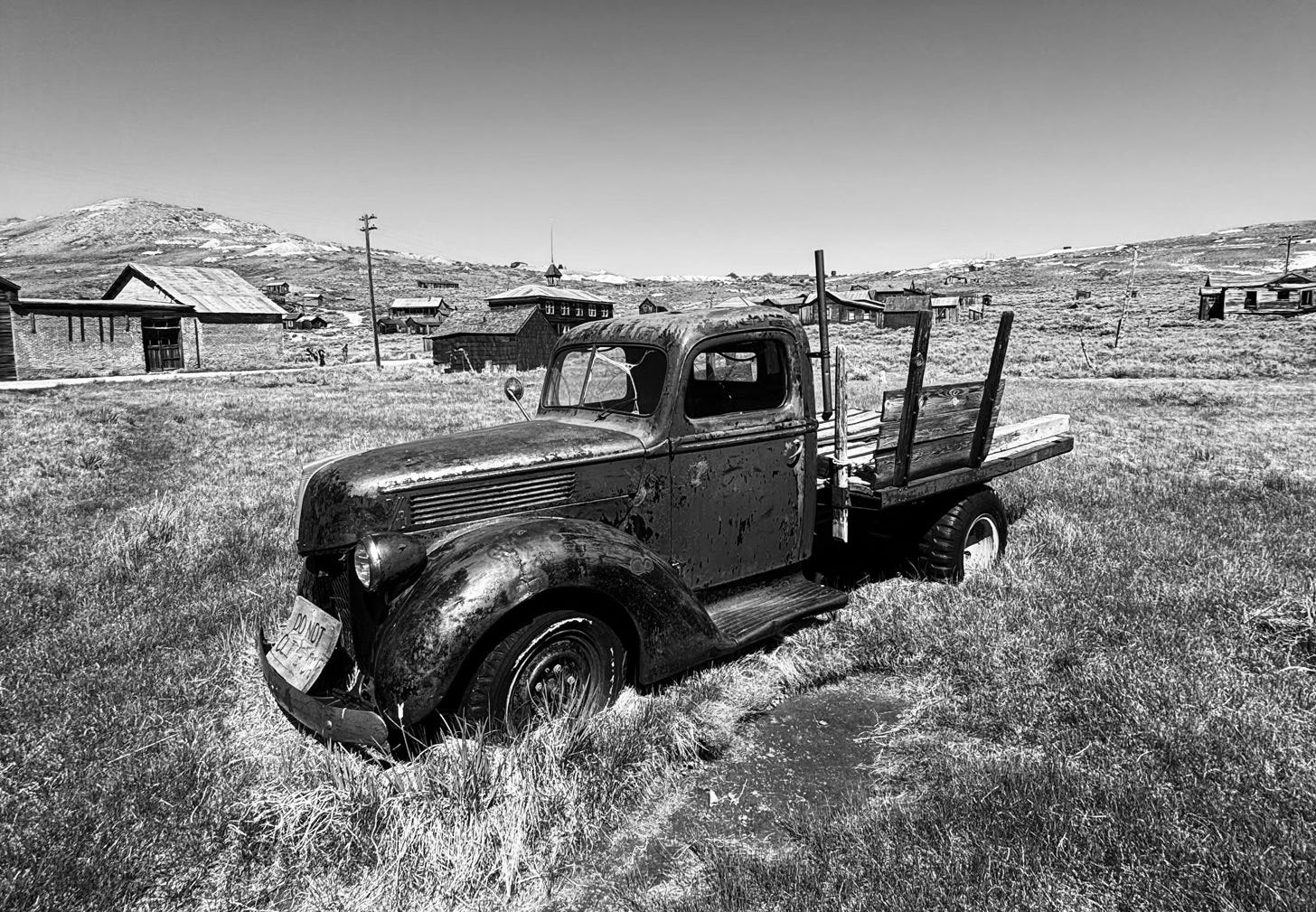 1940's car in decaying state sitting in field with abandoned buildings in background