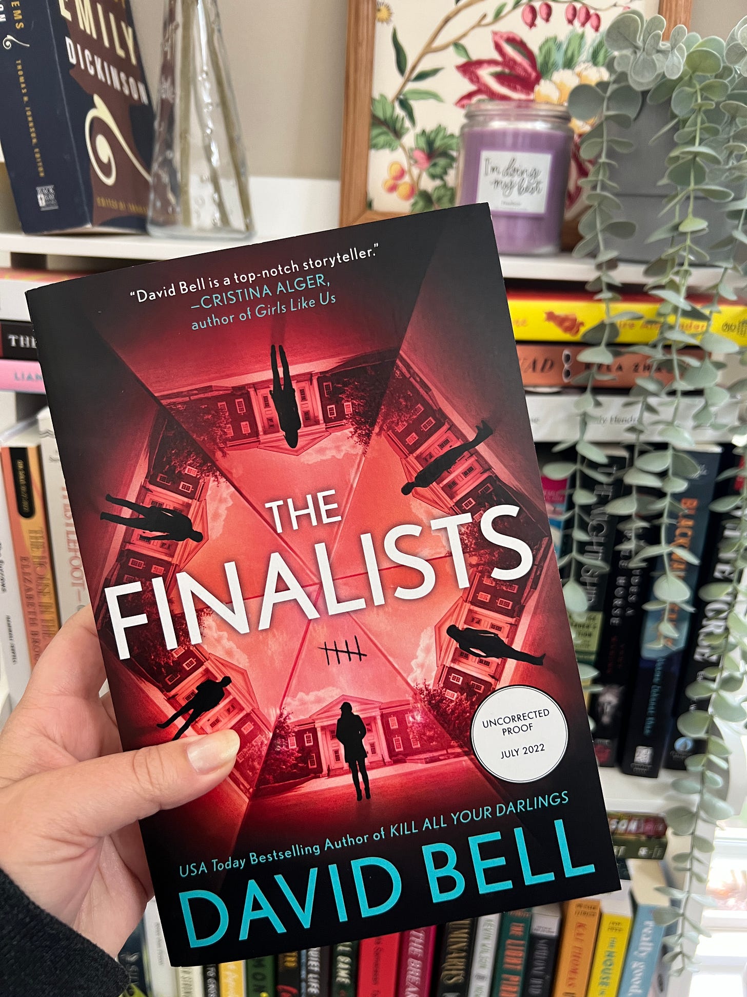A white hand holding an ARC of The Finalists up in front of a full bookshelf. A white hand holding an ARC of The Finalists up in front of a full bookshelf.