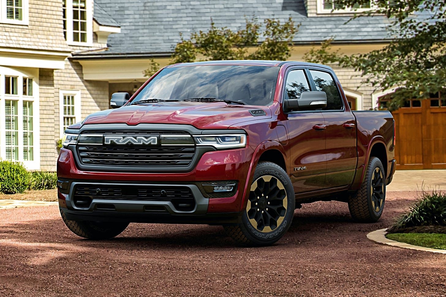 A red 2025 Ram Ramcharger electric truck is parked in the shade on a gravel driveway in front of a large tan home with a gray roof.