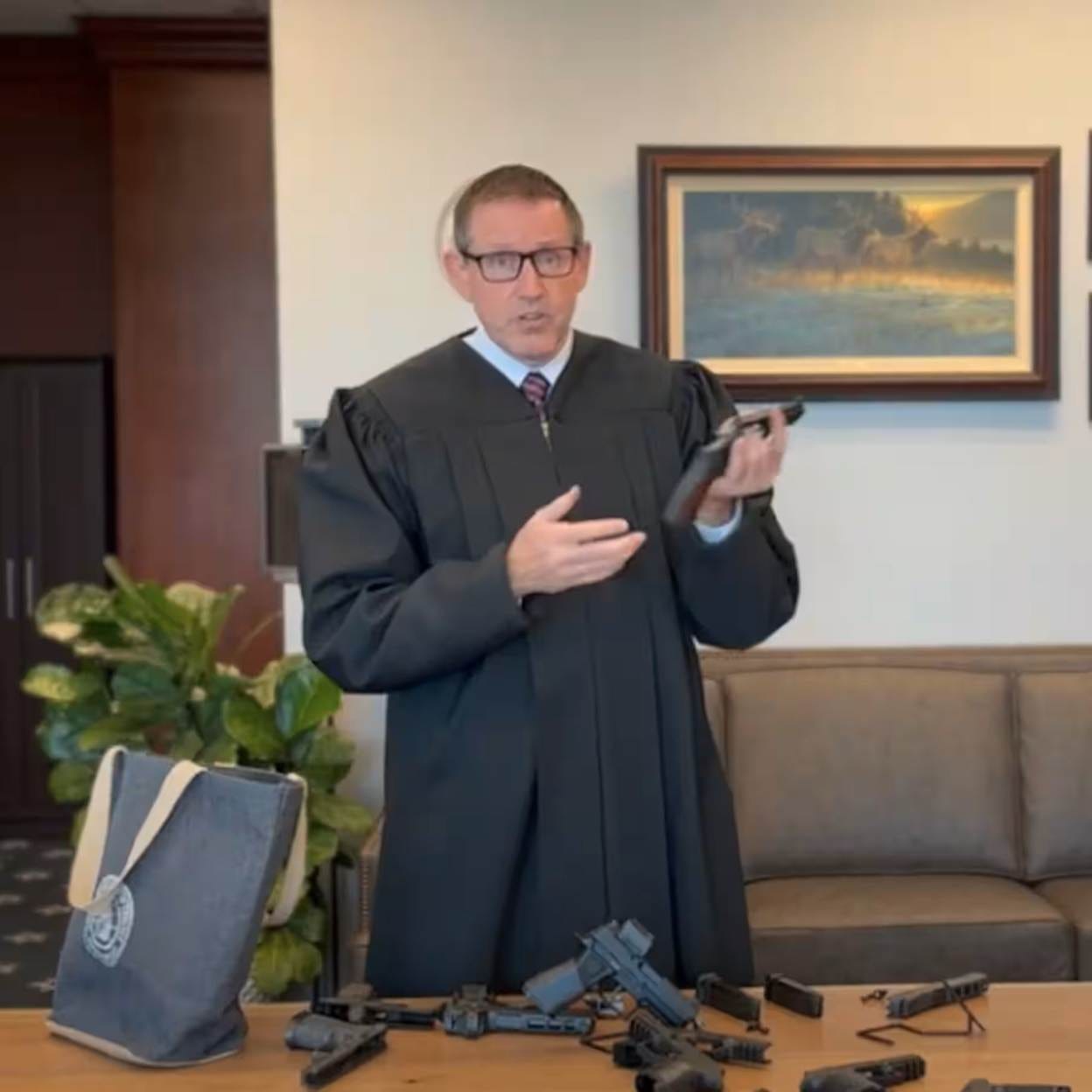 A lawyer in a black robe is holding a gun and a bag of ammunition in a room with a framed painting and a potted plant.

AI-generated content may be incorrect.