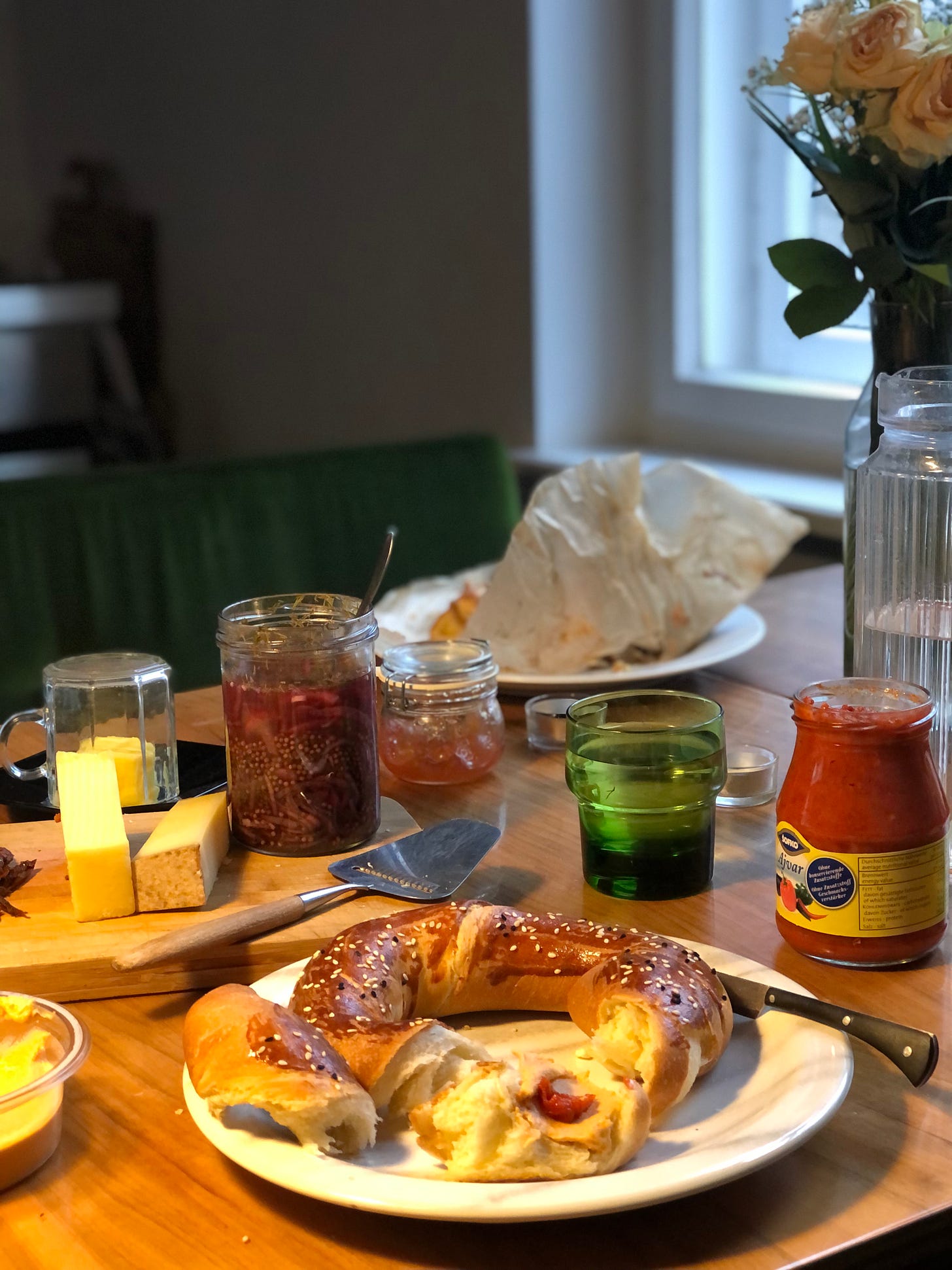 A view of a table with lots of items across it, lit by warm lamp light from the left and cool daylight from the right. In the foregroungd a plate with a ring shaped bread roll, behind which some cheese and conserve on a board behind. To the right, a cup, a jar of red something, a jug of water, a bunch of flowers. 