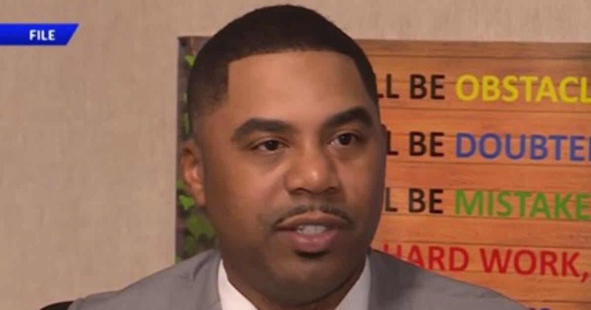 Man in a suit speaking during an interview, with motivational quotes visible in the background.