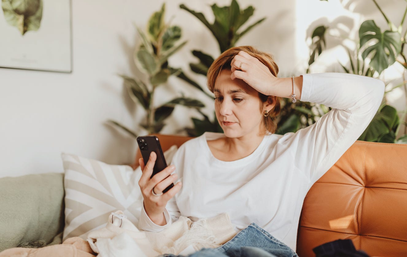Woman sitting on her couch with plants behind it. She is hesitantly looking at her phone. Woman sitting on her couch with plants behind it. She is hesitantly looking at her phone.