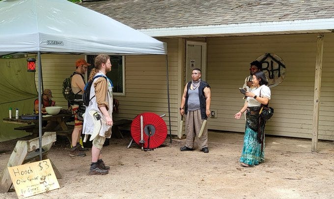 4 cowled and masked figures at night holding a finger over their mouth shushing the camera. A LARPer striking someone dressed as a zombie, exposed ribs and gorey makeup, with a foam sword. LARPers standing in the woods. One wears a torn lab coat and reinforced leather armor, and another in an orange baseball cap weilds a nerf gun and has too many tool belts. That's me, with the orange baseball cap. A third is mostly out of frame, but their large foam axe sticks up into view. A collection of LARPers stand outside a cabin with "RH" on the wall and a pop-up tent outside. Weapons and gear lay around the area and on the table.