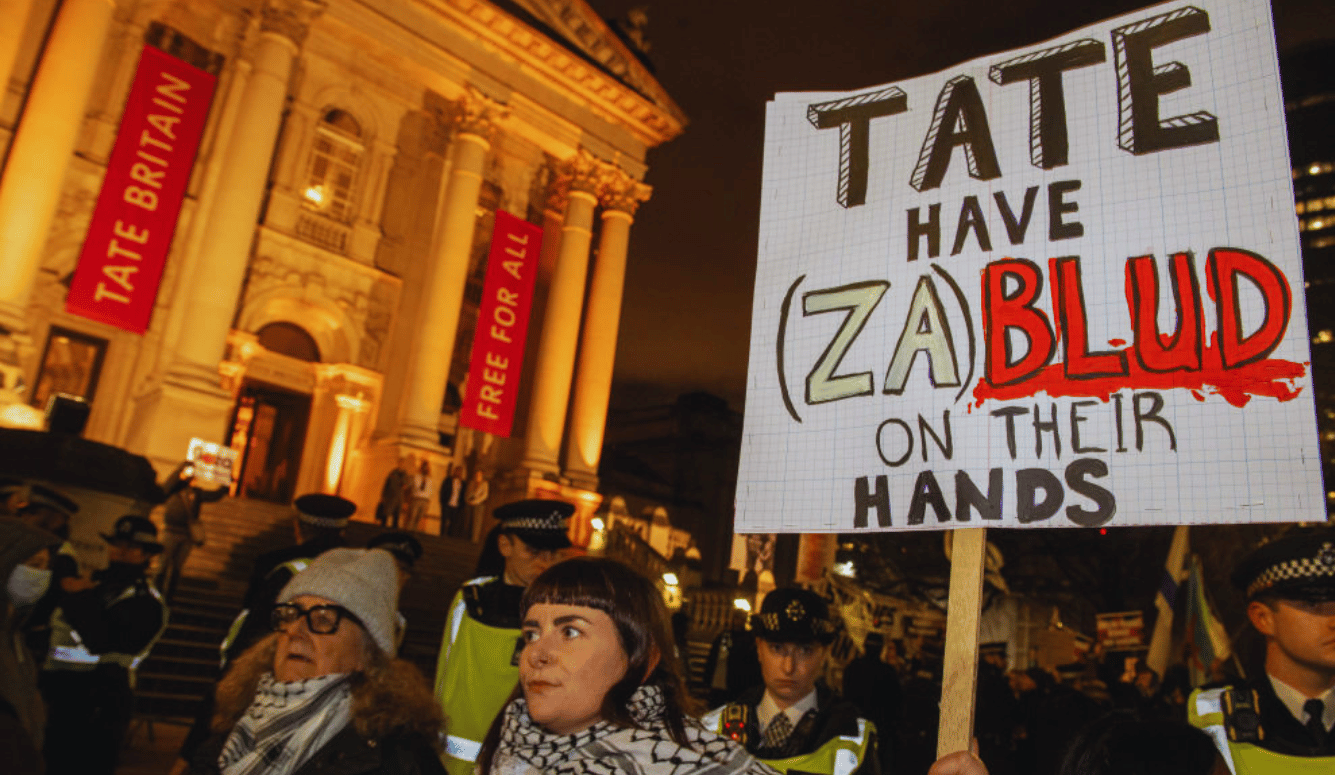 Protester holds a sign outside Tate Britain accusing the gallery of bloodshed as police and demonstrators stand nearby. Protester holds a sign outside Tate Britain accusing the gallery of bloodshed as police and demonstrators stand nearby.