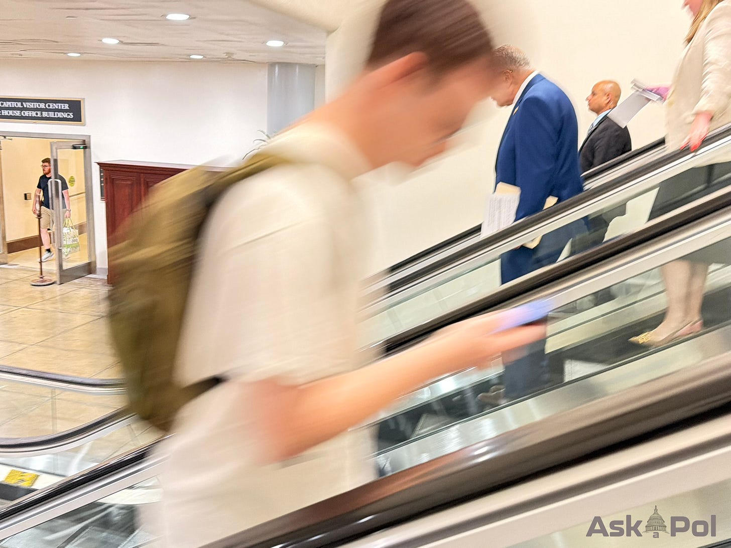A young man walks blurried in foreground as a politician rides an elevator down 10 or so feet in back of him A young man walks blurried in foreground as a politician rides an elevator down 10 or so feet in back of him