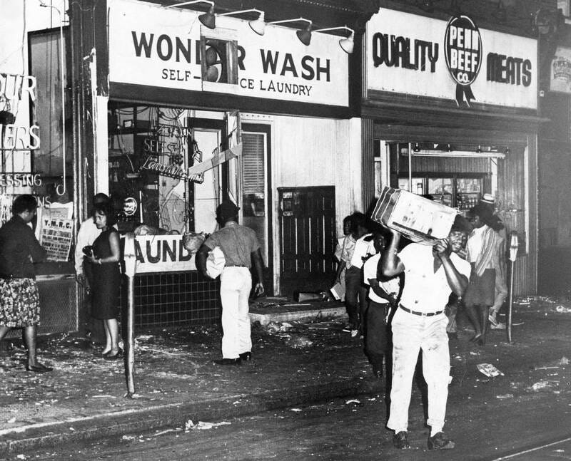 A Black man carries a large appliance out of a damaged "Wonder Wash Self-Service Laundry" storefront with broken windows, while debris litters the sidewalk.