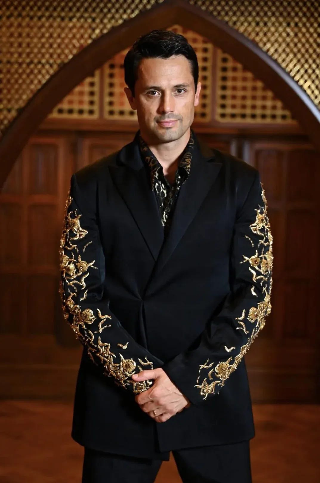 Stephen in a black suit jacket with gold floral embroidery on the sleeves, matching black and gold patterned shirt, hands clasped in front.