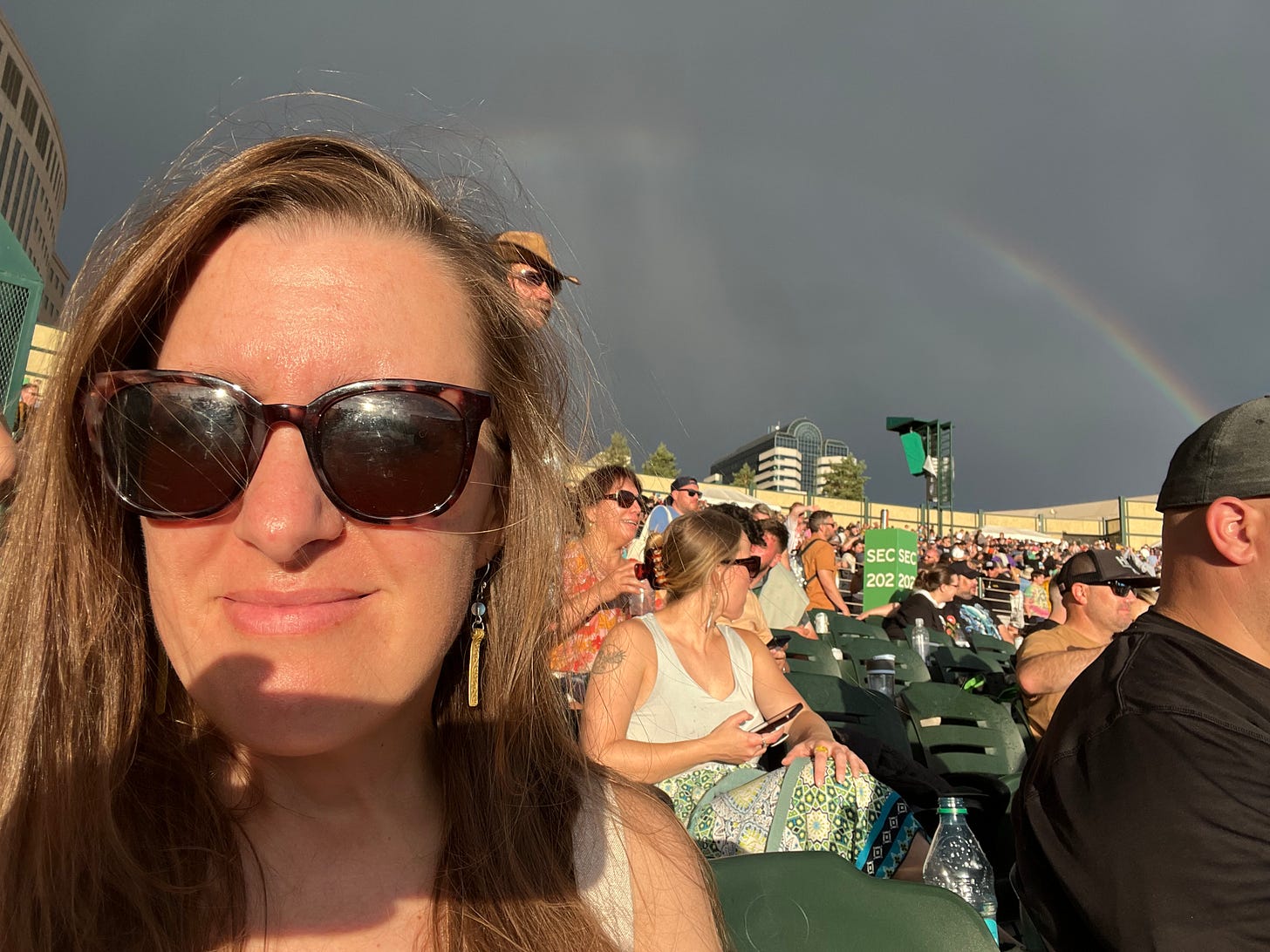 Amy in sunglasses sitting in the amphitheater with dark clouds and a rainbow behind her